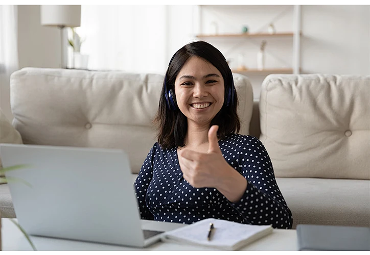 A woman wearing headphones, enjoying a laptop game and giving a thumbs-up.